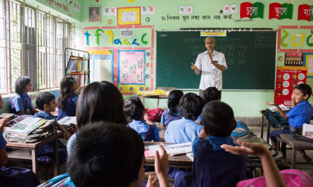 A male teacher stands at the front of a packed class of smiling students, Bangladesh.