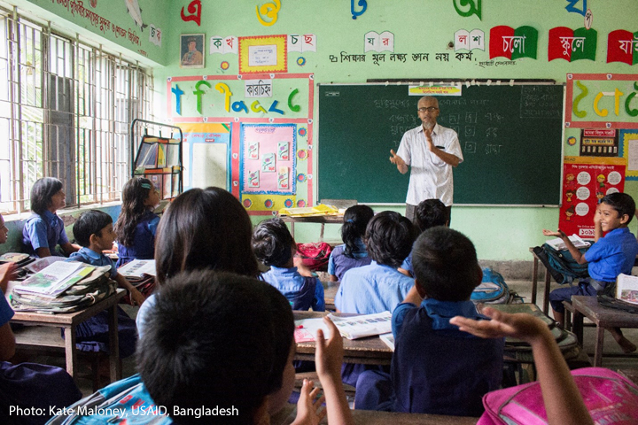 A male teacher stands at the front of a packed class of smiling students, Bangladesh.