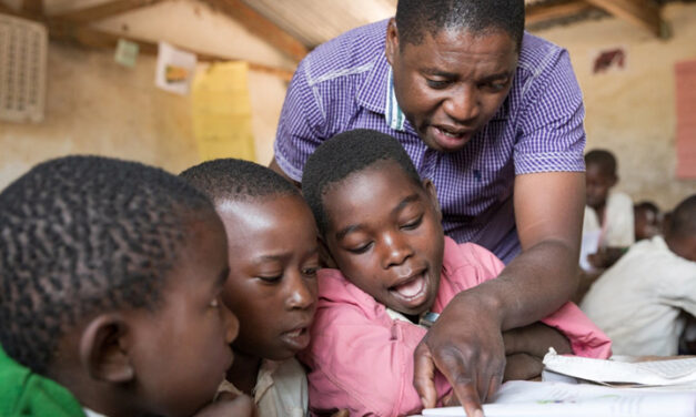 Students with teacher and textbook following her finger to read text at. Kasakola Primary School, Mpanda, Katavi, Tanzania.