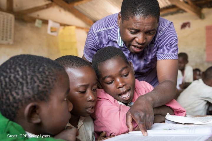 Students with teacher and textbook following her finger to read text at. Kasakola Primary School, Mpanda, Katavi, Tanzania.