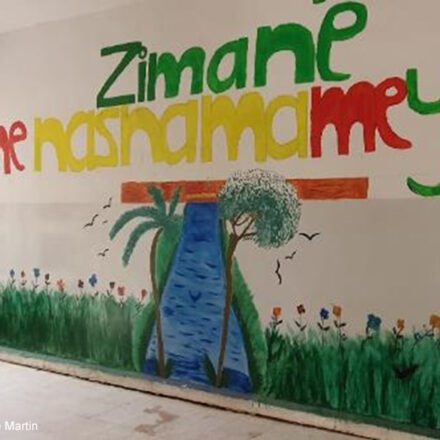 Mural on a wall in a Corridor in the school run by AANES in Qamishli. "Zimane me Nasnama me ye" with a meadow and a stream with trees.
