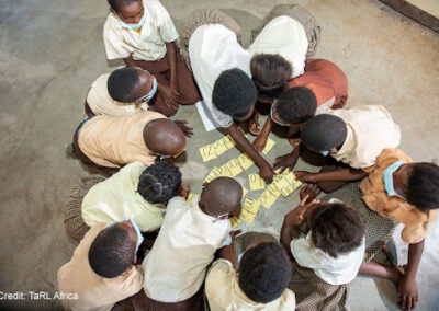 A group of children sit on the floor and gather around some cards with words to make a sentence.