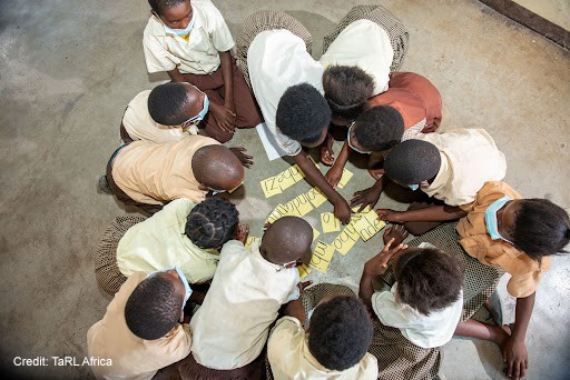 A group of children sit on the floor and gather around some cards with words to make a sentence.