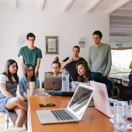 A group of students stand around laptops doing group work.