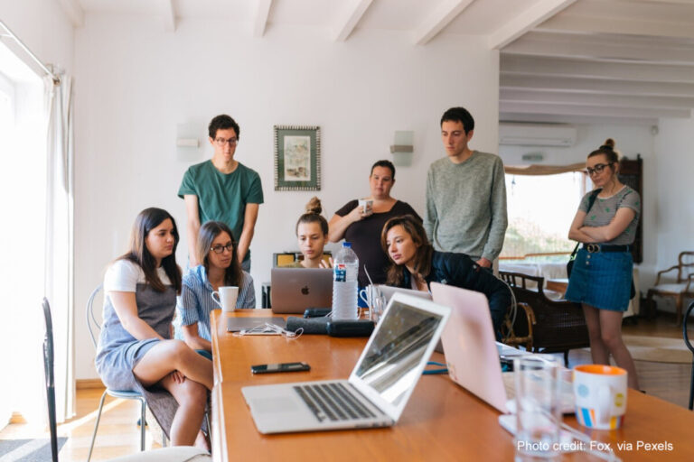 A group of students stand around laptops doing group work.
