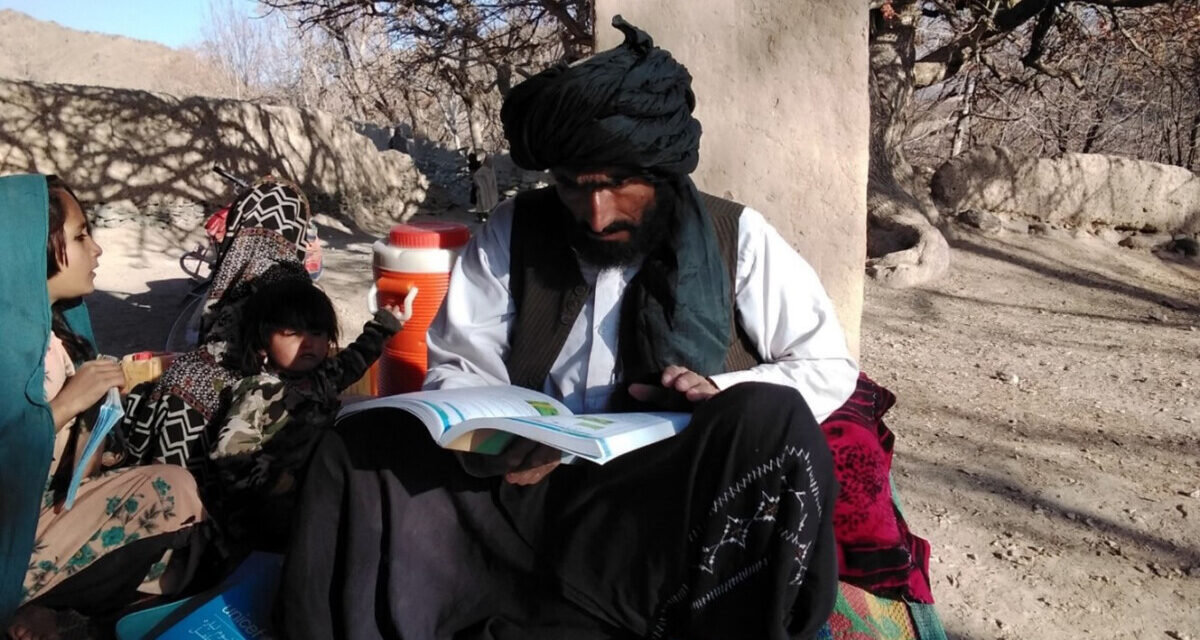 A male teacher sits on a rug outside with a teacher guide and young children next to him, Afghanistan.