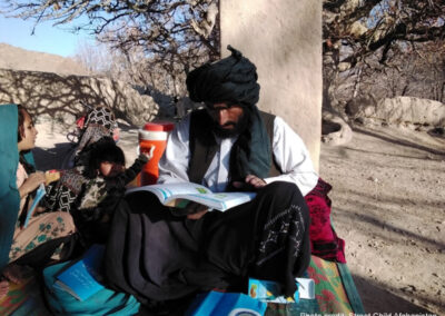 A male teacher sits on a rug outside with a teacher guide and young children next to him, Afghanistan.