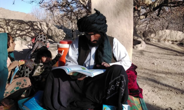 A male teacher sits on a rug outside with a teacher guide and young children next to him, Afghanistan.