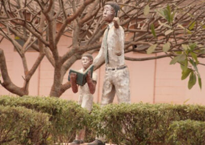 Statue of a teacher and a child with a book. Statue is set in amongst trees and shrubs