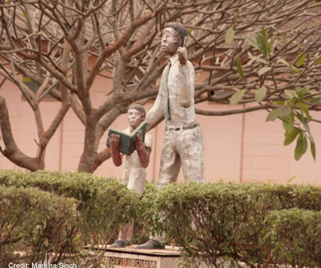 Statue of a teacher and a child with a book. Statue is set in amongst trees and shrubs