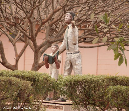 Statue of a teacher and a child with a book. Statue is set in amongst trees and shrubs