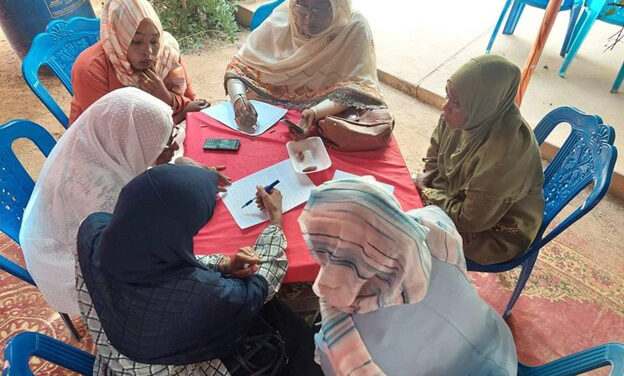 A group of women sit around a table working together on peacebuilding awareness plans.