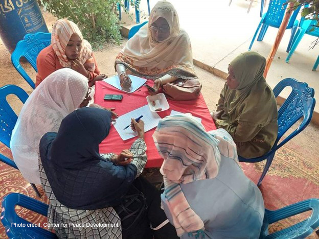 A group of women sit around a table working together on peacebuilding awareness plans.