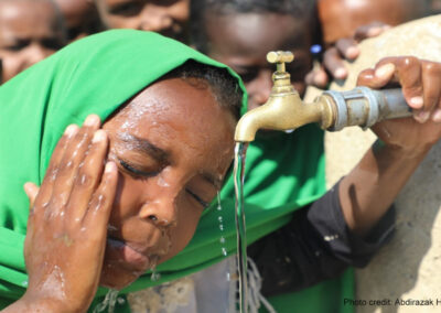 a young child splashes water on her face from the school tap, with other children waiting their turn behind her.
