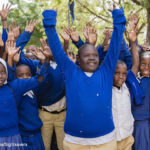 Isaya at school with his peers in Tanzania. Children in blue uniform jumpers holding their hands in the air in a group.