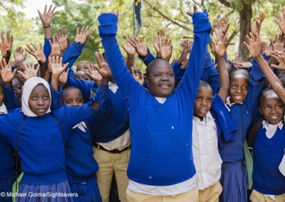 Isaya at school with his peers in Tanzania. Children in blue uniform jumpers holding their hands in the air in a group.