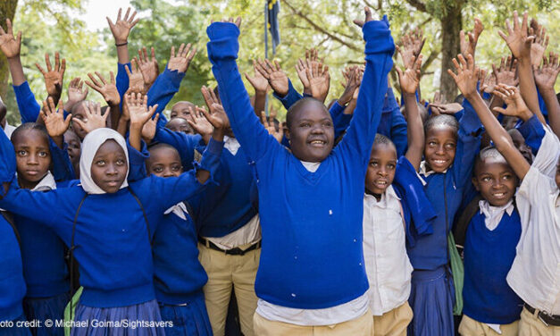Isaya at school with his peers in Tanzania. Children in blue uniform jumpers holding their hands in the air in a group.