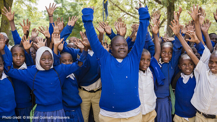 Isaya at school with his peers in Tanzania. Children in blue uniform jumpers holding their hands in the air in a group.