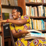 Woman sitting in a library in Ghana. Selecting a book from shelves with some on her knee.