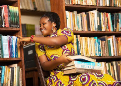 Woman sitting in a library in Ghana. Selecting a book from shelves with some on her knee.