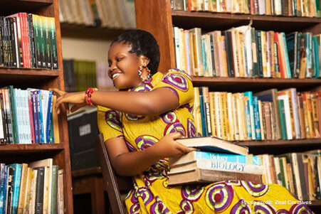 Woman sitting in a library in Ghana. Selecting a book from shelves with some on her knee.