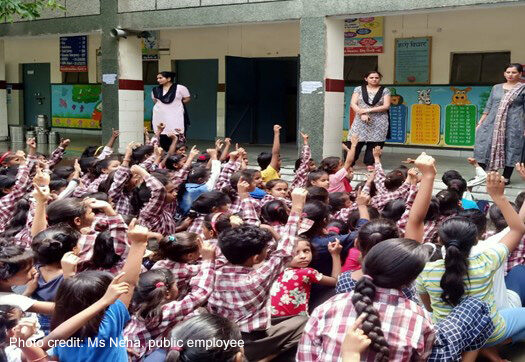 Teachers stand at the front of an outdoor class of primary students, India