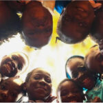 Children stand in a circle and look down to the camera in the middle of the floor.