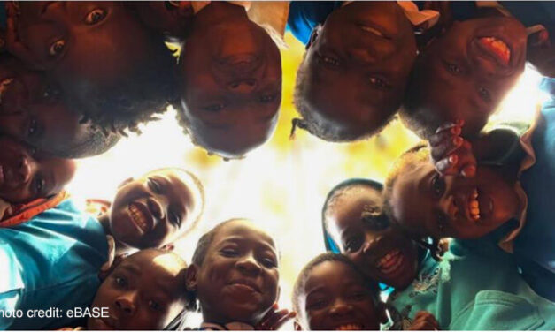 Children stand in a circle and look down to the camera in the middle of the floor.