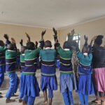 Children take part in a learning activity jumping in a circle with hands above their heads, around the classroom, Tanzania.