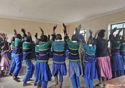 Children take part in a learning activity jumping in a circle with hands above their heads, around the classroom, Tanzania.