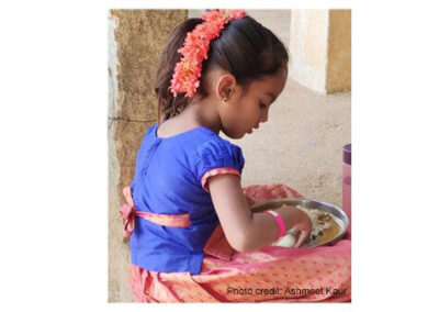 A young girl sits cross-legged with a plate of food on her lap, India.