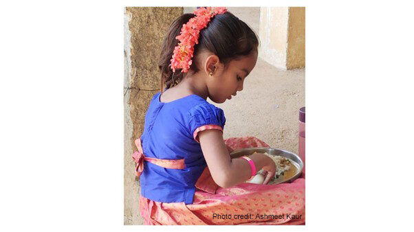 A young girl sits cross-legged with a plate of food on her lap, India.