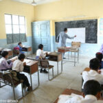 Male teacher writes on the blackboard in class with students sitting at their desks writing, Pakistan.