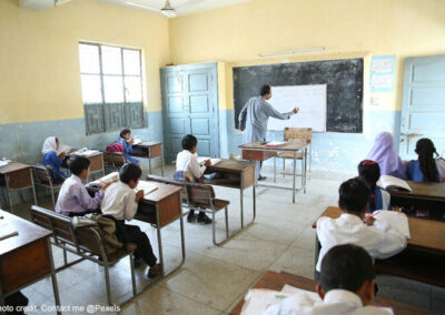 Male teacher writes on the blackboard in class with students sitting at their desks writing, Pakistan.