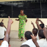 Teacher Amunazo Belinda, wearing a green costume standing at the front of a classroom of children with raised hands, Democratic Republic of Congo.