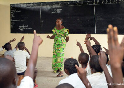 Teacher Amunazo Belinda, wearing a green costume standing at the front of a classroom of children with raised hands, Democratic Republic of Congo.