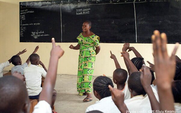 Teacher Amunazo Belinda, wearing a green costume standing at the front of a classroom of children with raised hands, Democratic Republic of Congo.