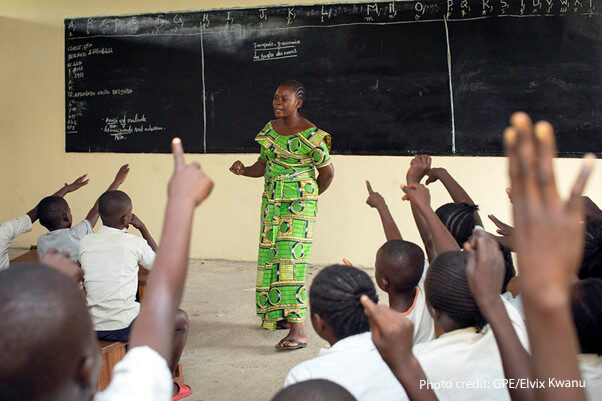 Teacher Amunazo Belinda, wearing a green costume standing at the front of a classroom of children with raised hands, Democratic Republic of Congo.