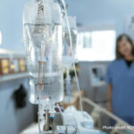 A nurse stands talking to a patient in a hospital bed in the blurred background. In the fore is an IV drip on a stand.