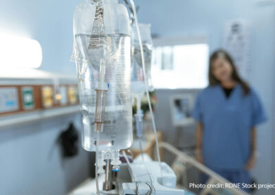 A nurse stands talking to a patient in a hospital bed in the blurred background. In the fore is an IV drip on a stand.