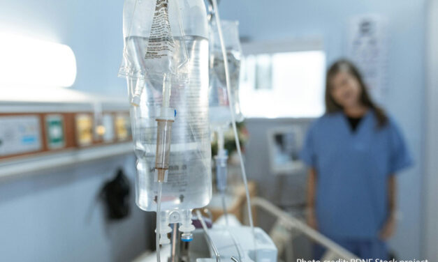 A nurse stands talking to a patient in a hospital bed in the blurred background. In the fore is an IV drip on a stand.