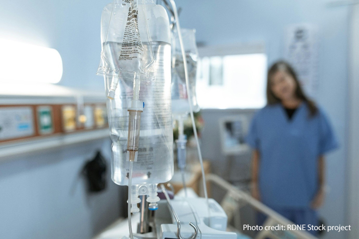 A nurse stands talking to a patient in a hospital bed in the blurred background. In the fore is an IV drip on a stand.