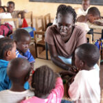 Nyuriel, a learning support assistant is teaching a group of children sitting around a table in a classroom in Kakuma refugee camp in Kenya.