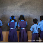 Five girls and boys stand facing the blackboard in a classroom, India.