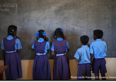 Five girls and boys stand facing the blackboard in a classroom, India.