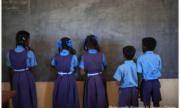 Five girls and boys stand facing the blackboard in a classroom, India.