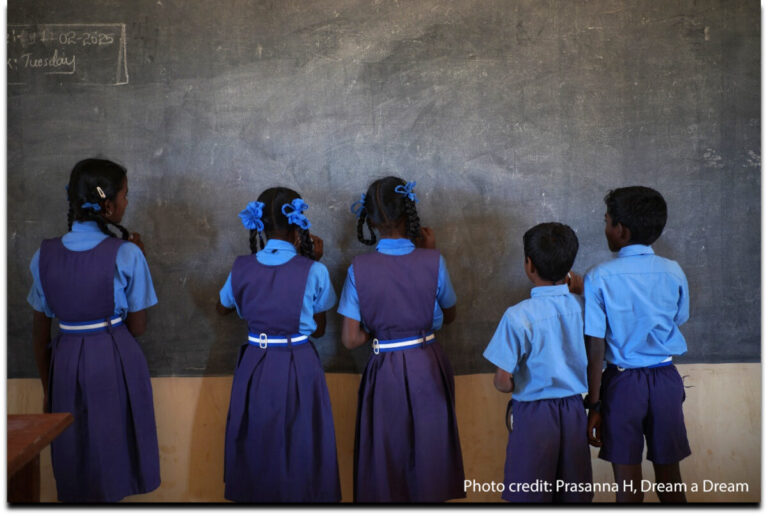 Five girls and boys stand facing the blackboard in a classroom, India.