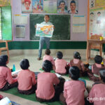 A male teacher stands at the front of a class with students sitting on mats on the floor, India.