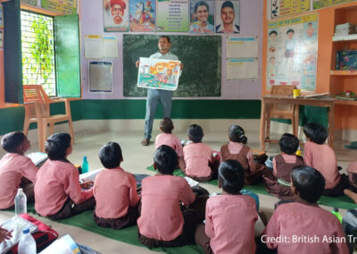 A male teacher stands at the front of a class with students sitting on mats on the floor, India.