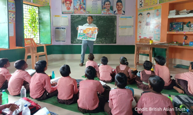 A male teacher stands at the front of a class with students sitting on mats on the floor, India.